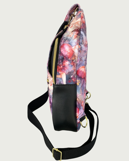 Handcrafted sling bag standing sideways with a print of an enchanted forest with fairies and mushrooms in pinks and purples, grays and oranges. Water-resistant canvas bag with black faux leather accents with an adjustable wide black cotton webbing strap also repositional for left and right handed wear. All hardware is gold including the zipper teeth on black zipper tape. A front panel that unhooks with a swivel held onto a d-ring is shown. Behind that is a curved zippered main compartment of black vinyl.