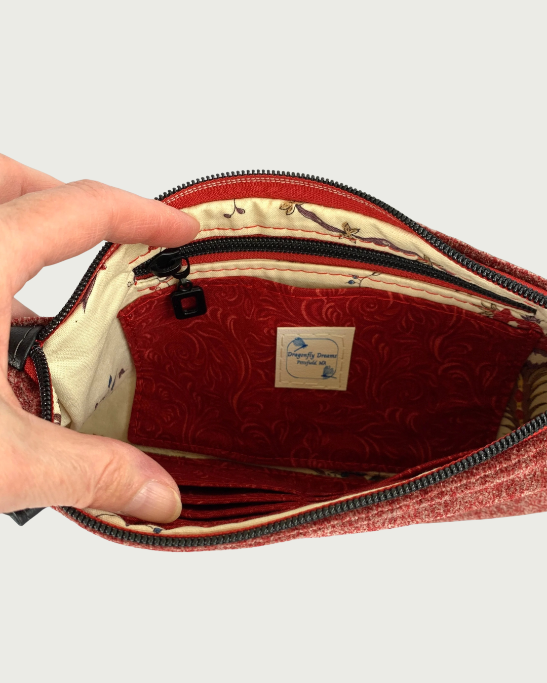 Handcrafted merlot chenille-like polyester wristlet wallet held open by a hand revealing a slip pocket and zippered pocket with a Dragonfly Dreams label sewn in. The pocket and main zipper is matt black hardware including an open square zipper pull and burgundy zipper tape. Partial buit-in card slots can be seen on the other side of the bag’s interior.