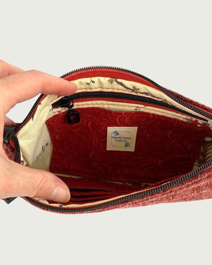 Handcrafted merlot chenille-like polyester wristlet wallet held open by a hand revealing a slip pocket and zippered pocket with a Dragonfly Dreams label sewn in. The pocket and main zipper is matt black hardware including an open square zipper pull and burgundy zipper tape. Partial buit-in card slots can be seen on the other side of the bag’s interior.