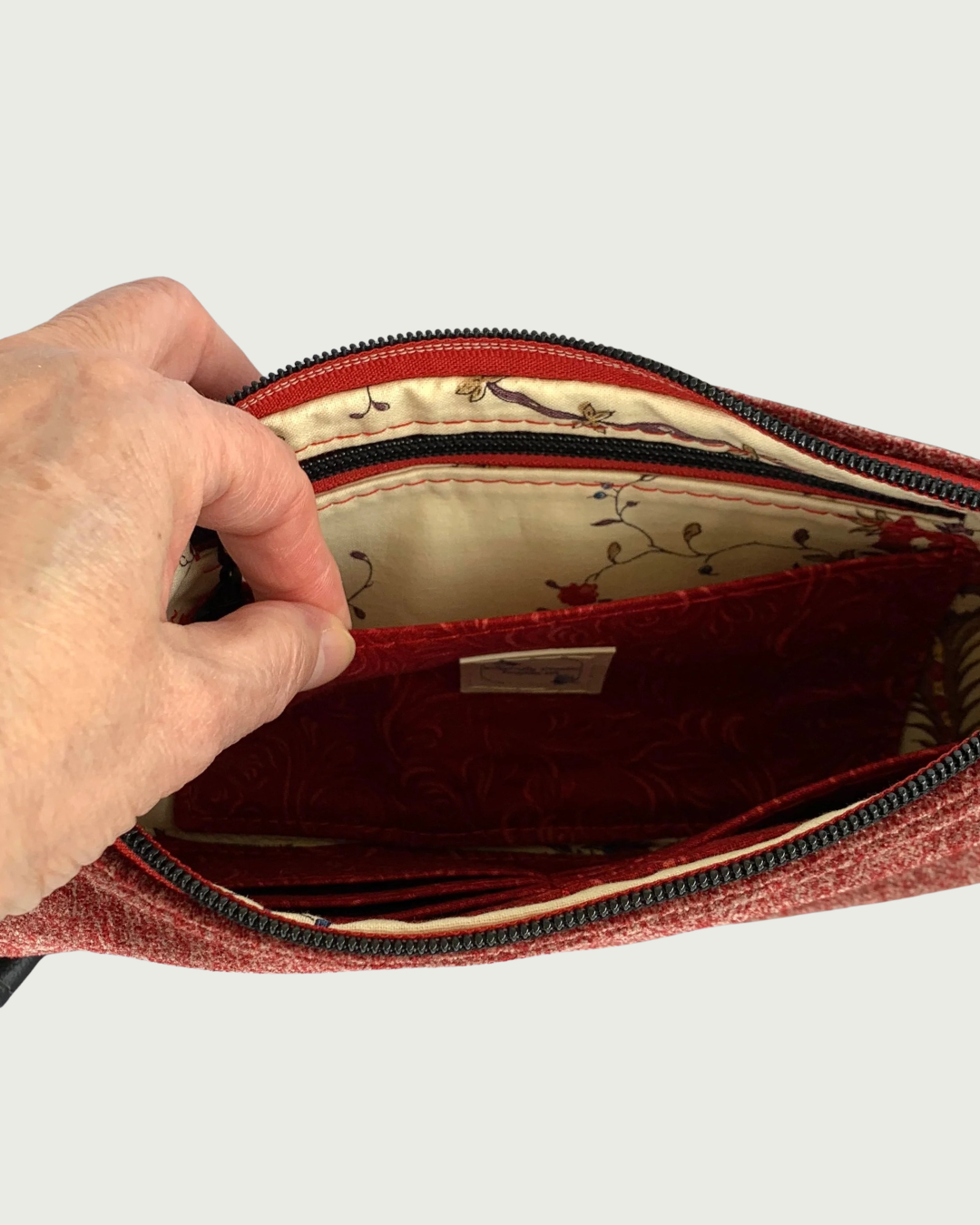 The interior of a handcrafted merlot chenille-like polyester wristlet wallet with a hand holding open a slip pocket in front of a zippered pocket with a Dragonfly Dreams label sewn in. The pocket and main zipper is matt black hardware and the zipper tape is burgundy. Partial buit-in card slots can be seen on the other side of the bag’s interior.