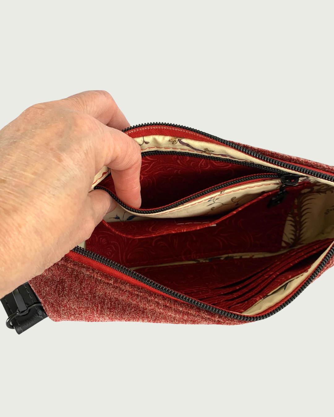 The interior of a handcrafted merlot chenille-like polyester wristlet wallet with fingers holding open both a slip pocket and a zippered pocket behind it. The pocket and main zipper is matt black hardware and the zipper tape is burgundy. Partial buit-in card slots can be seen on the other side of the bag’s interior.