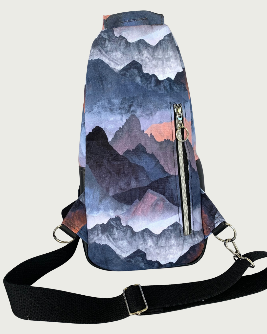 Back view of handcrafted sling bag with a print of mountains in blues, grays, and corals. Water-resistant canvas bag with an adjustable wide black cotton webbing strap also repositional for left and right handed wear. All hardware is nickel including the zipper teeth on black zipper tape that is positioned vertically for a concealed pocket.