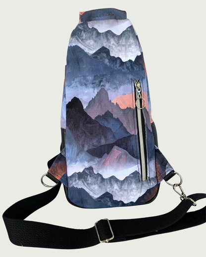 Back view of handcrafted sling bag with a print of mountains in blues, grays, and corals. Water-resistant canvas bag with an adjustable wide black cotton webbing strap also repositional for left and right handed wear. All hardware is nickel including the zipper teeth on black zipper tape that is positioned vertically for a concealed pocket.