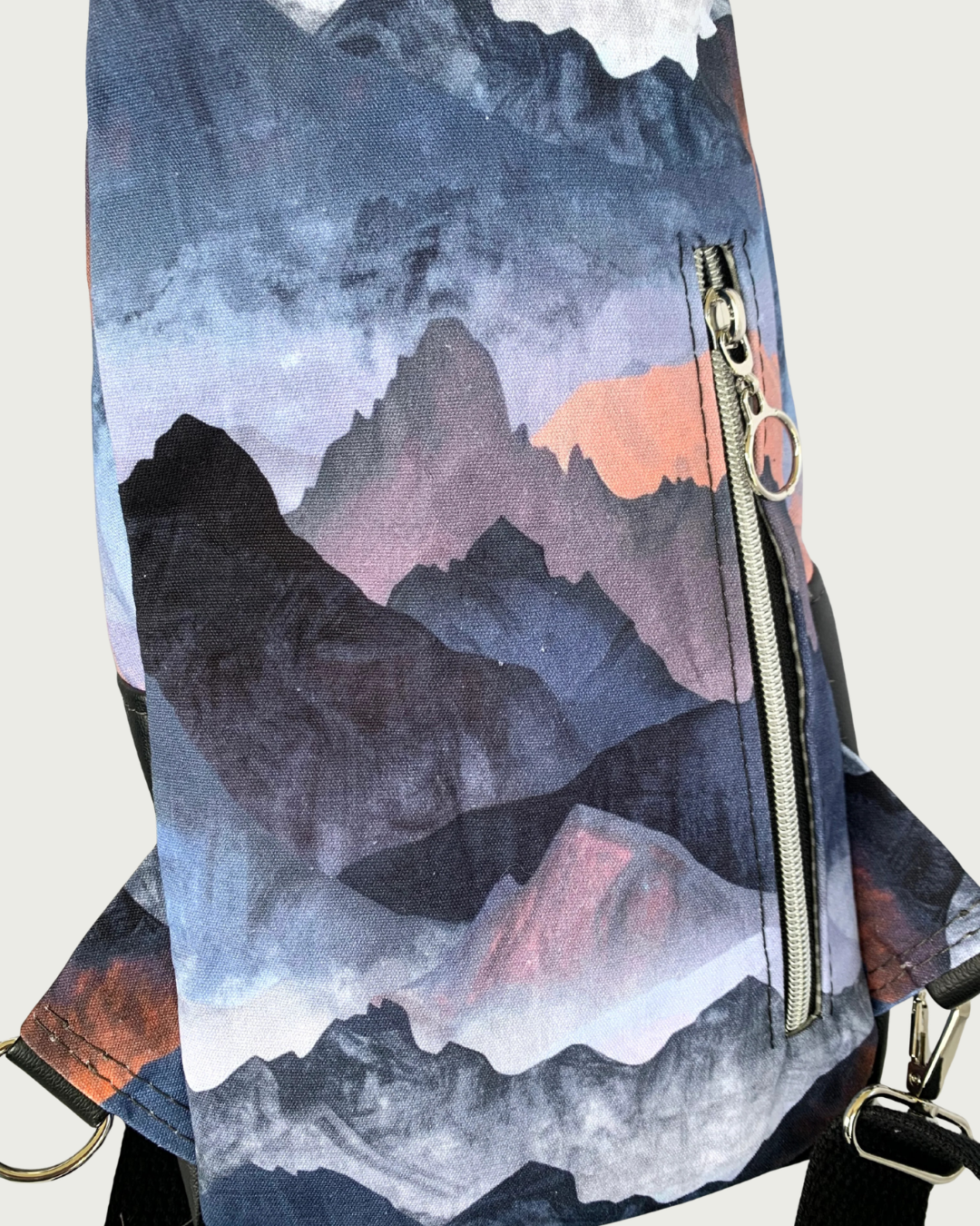 Back view of handcrafted sling bag with a print of mountains in blues, grays, and corals. Water-resistant canvas bag with an adjustable wide black cotton webbing strap also repositional for left and right handed wear. All hardware is nickel including the zipper teeth on black zipper tape that is positioned vertically for a concealed pocket.