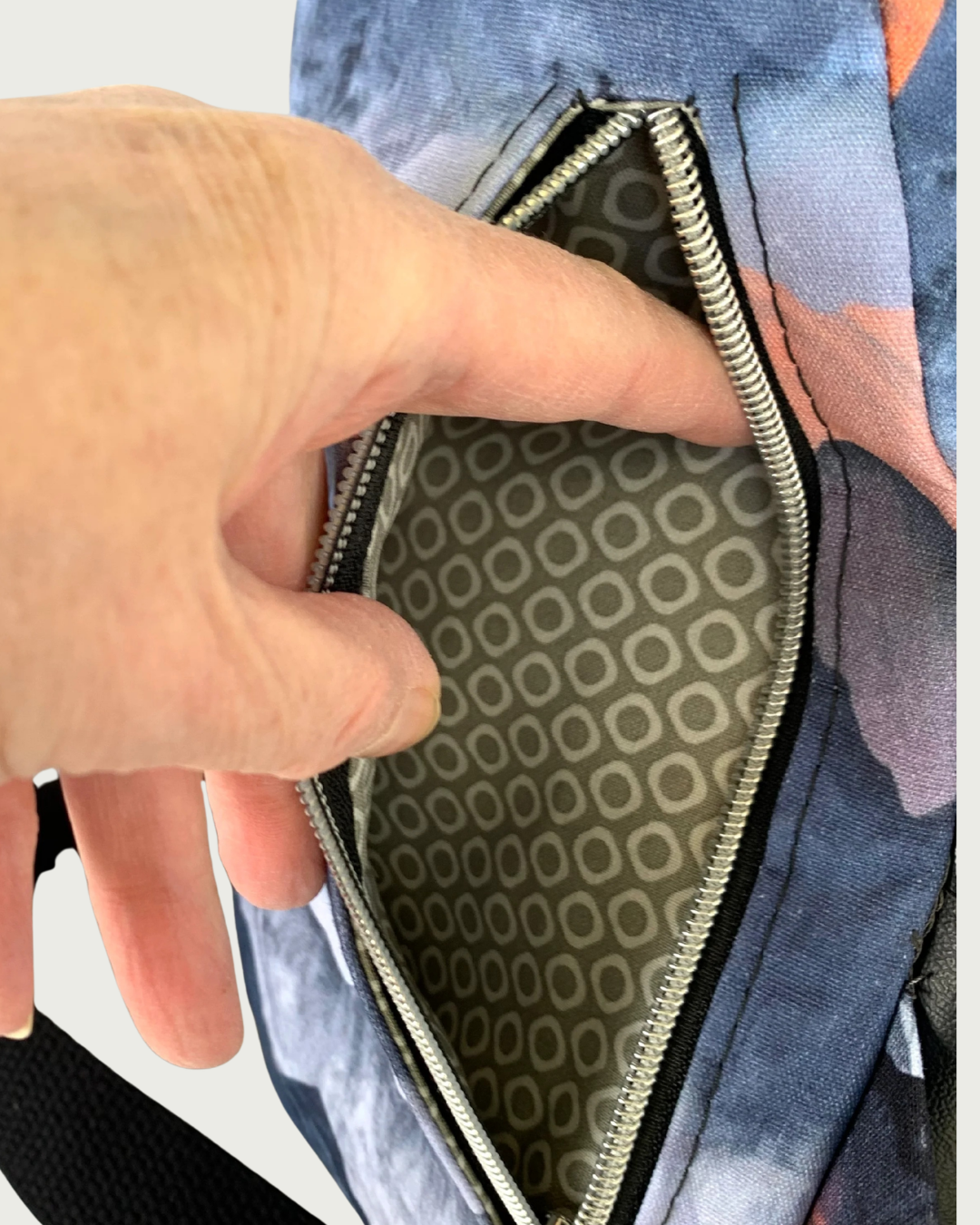 Back view of handcrafted sling bag of water-resistant canvas in blues, grays, and corals. A hand is holding open the back vertical zippered pocket. The pocket lining is a light gray with a white cheerios pattern.