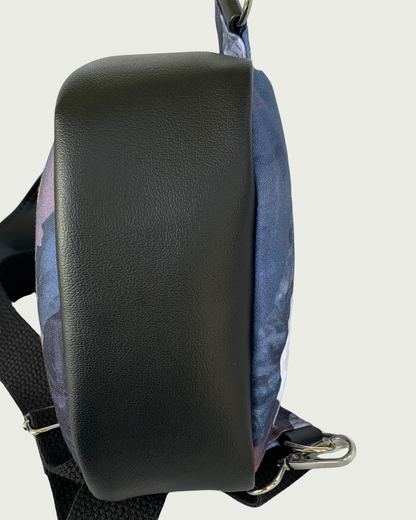 The bottom view of a handcrafted sling bag of water-resistant canvas in blues, grays, and corals. The bottom is also part of the gusset and it is made from black faux leather. An adjustable wide black cotton webbing strap is shown. All hardware is nickel.