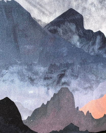 A closeup view of the fabric of a sling bag of water-resistant canvas with a print of mountains in blues, grays, and corals. This is a closeup view between the side black vinyl gusset and the front zippered main compartment and zippered pocket. All hardware is nickel including the zipper teeth on black zipper tape with circle zipper pulls.