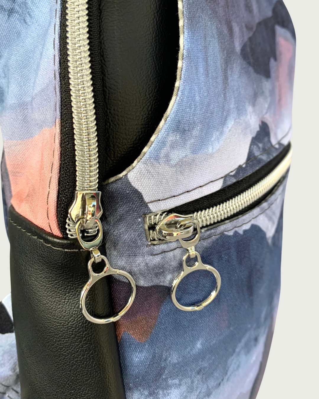 A handcrafted sling bag of water-resistant canvas with a print of mountains in blues, grays, and corals. This is a closeup view between the side black vinyl gusset and the front zippered main compartment and zippered pocket. All hardware is nickel including the zipper teeth on black zipper tape with circle zipper pulls.