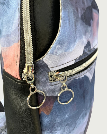 A handcrafted sling bag of water-resistant canvas with a print of mountains in blues, grays, and corals. This is a closeup view between the side black vinyl gusset and the front zippered main compartment and zippered pocket. All hardware is nickel including the zipper teeth on black zipper tape with circle zipper pulls.