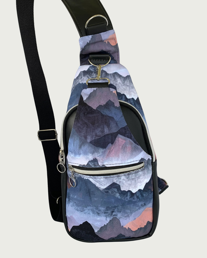 Handcrafted sling bag with a print of mountains in blues, grays, and corals. Water-resistant canvas bag with black faux leather accents with an adjustable wide black cotton webbing strap also repositional for left and right handed wear. All hardware is nickel including the zipper teeth on black zipper tape. A front zipper pocket is shown as well as a front panel that unhooks with a swivel held onto a d-ring. Behind that is a curved zippered main compartment.