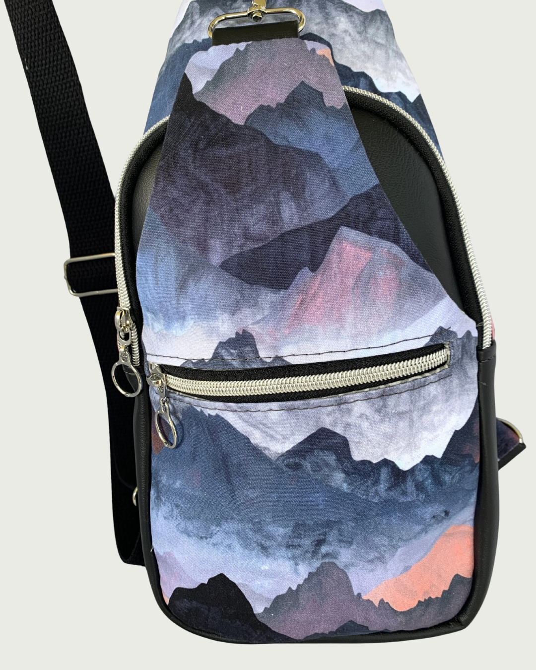 Handcrafted Sling bag with a print of mountains in blues, grays, and corals. Water Resistant canvas bag with black faux leather accents with an adjustable wide black cotton webbing strap also repositional for left and right handed wear. All hardware is nickel including the zipper teeth on black zipper tape. A front zipper pocket is shown as well as a front panel that unhooks with a swivel held onto a d-ring. Behind that is a curved zippered main compartment.
