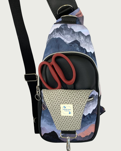 Handcrafted sling bag with a print of mountains in blues, grays, and corals. Water-resistant canvas bag with black faux leather accents with an adjustable wide black cotton webbing strap. All hardware is nickel including the zipper teeth on black zipper tape. A front zipper pocket is partially shown on an open panel holding red-handled scissors inside. Behind that is the main compartment with a curved zipper. The lining of the front panel is a light gray with a white cheerios pattern. A Dragonfly Dreams lab