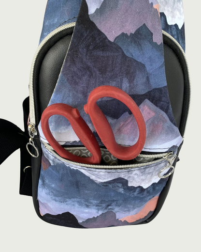 Handcrafted sling bag with a print of mountains in blues, grays, and corals. Water-resistant canvas bag with black faux leather accents with an adjustable wide black cotton webbing strap. All hardware is nickel including the zipper teeth on black zipper tape. A front zipper pocket is shown open with red-handled scissors inside. Behind that is the main compartment with a curved zipper.