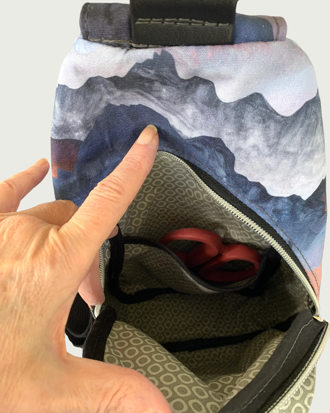 Handcrafted sling bag with a print of mountains in blues, grays, and corals on water-resistant canvas. A hand is holding open the main compartment revealing a lining of light gray and white cheerios pattern with black binding on the seams. The interior slip pocket has a pair of red-handled scissors peeking through.