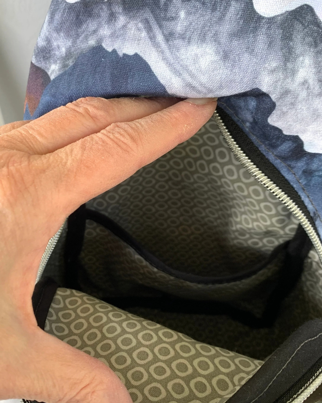 Handcrafted sling bag with a print of mountains in blues, grays, and corals on water-resistant canvas. A hand is holding open the main compartment revealing a lining of light gray and white cheerios pattern, a slip pocket, and black binding on the seams.