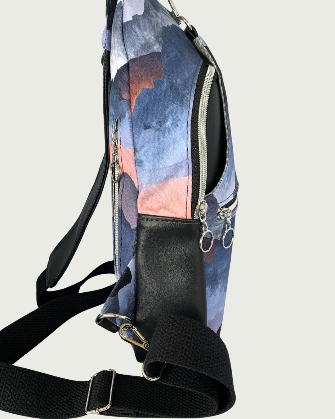 Handcrafted sling bag with a print of mountains in blues, grays, and corals standing sideways. Water-resistant canvas bag with black faux leather accents with an adjustable wide black cotton webbing strap. All hardware is nickel including the zipper teeth on black zipper tape on the front and back pockets as well as the main compartment. A front panel that has the pocket unhooks with a swivel held onto a d-ring. Behind that is a curved zippered main compartment made of the black vinyl.