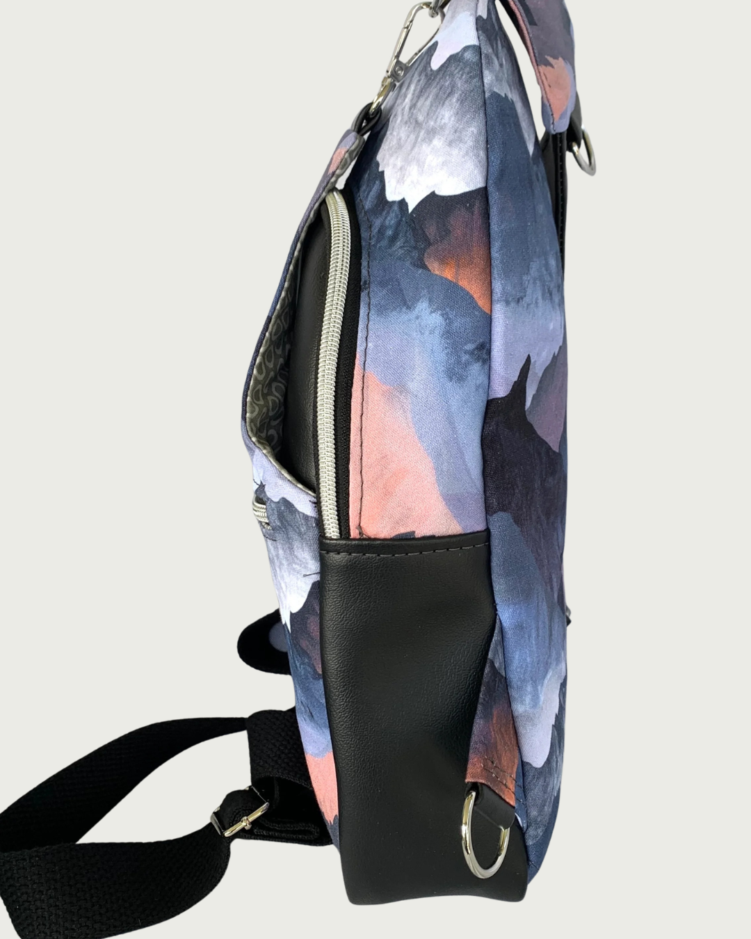 Handcrafted sling bag with a print of mountains in blues, grays, and corals standing sideways. Water-resistant canvas bag with black faux leather accents with an adjustable wide black cotton webbing strap also repositional for left and right handed wear. All hardware is nickel including the zipper teeth on black zipper tape. A front zipper pocket is shown as well as a front panel that unhooks with a swivel held onto a d-ring. Behind that is a curved zippered main compartment.