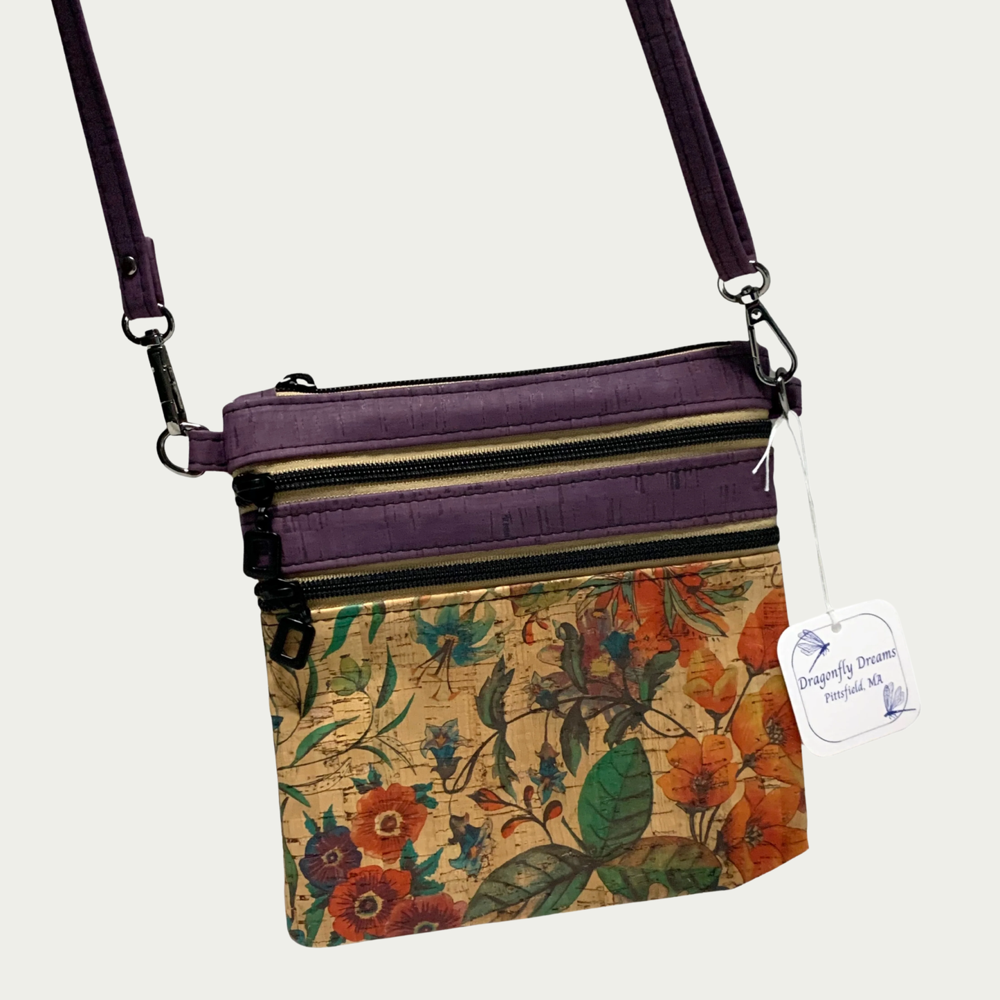 A crossbody bag of multicolored floral patterned cork with solid purple cork accents, two zippered pockets, a top zippered closure and matt black hardware. The adjustable, detachable strap is solid purple cork.