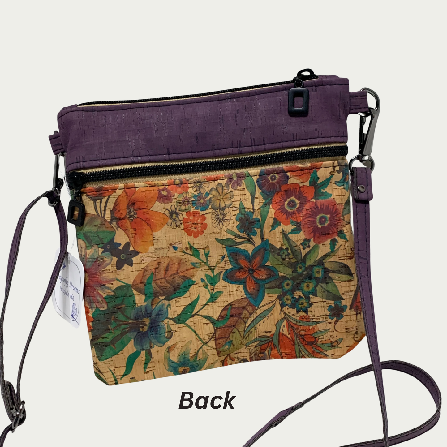 A crossbody bag of multicolored floral patterned cork with solid purple cork accents, a zippered pocket and a top zippered closure and matt black hardware. The adjustable, detachable strap is solid purple cork.