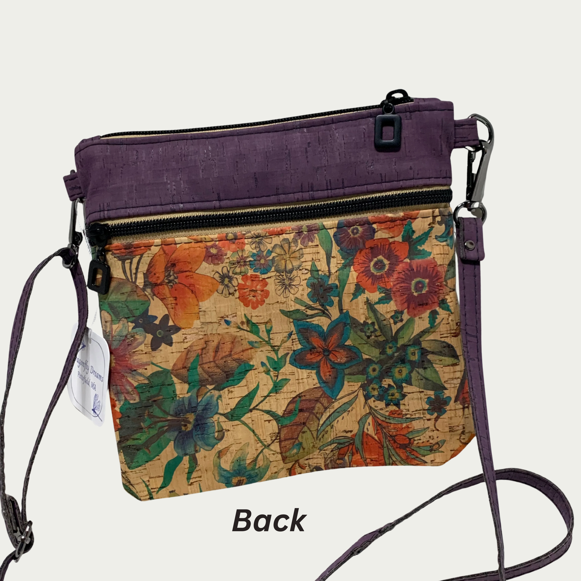 A crossbody bag of multicolored floral patterned cork with solid purple cork accents, a zippered pocket and a top zippered closure and matt black hardware. The adjustable, detachable strap is solid purple cork.