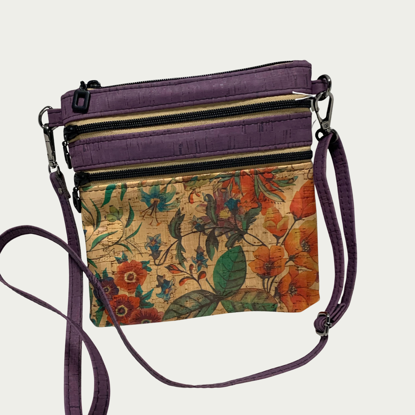 A crossbody bag of multicolored floral patterned cork with solid purple cork accents, two zippered pockets and a top zippered closure. The adjustable, detachable strap is solid purple cork.