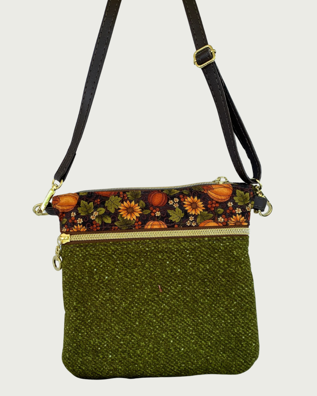Handmade olive green crossbody bag with a textured fabric body, a zippered pocket featuring a pumpkin and sunflower patterned band between above it, gold-tone hardware, a top zippered closure, hanging by a thin, coffee adjustable and detachable shoulder strap.
