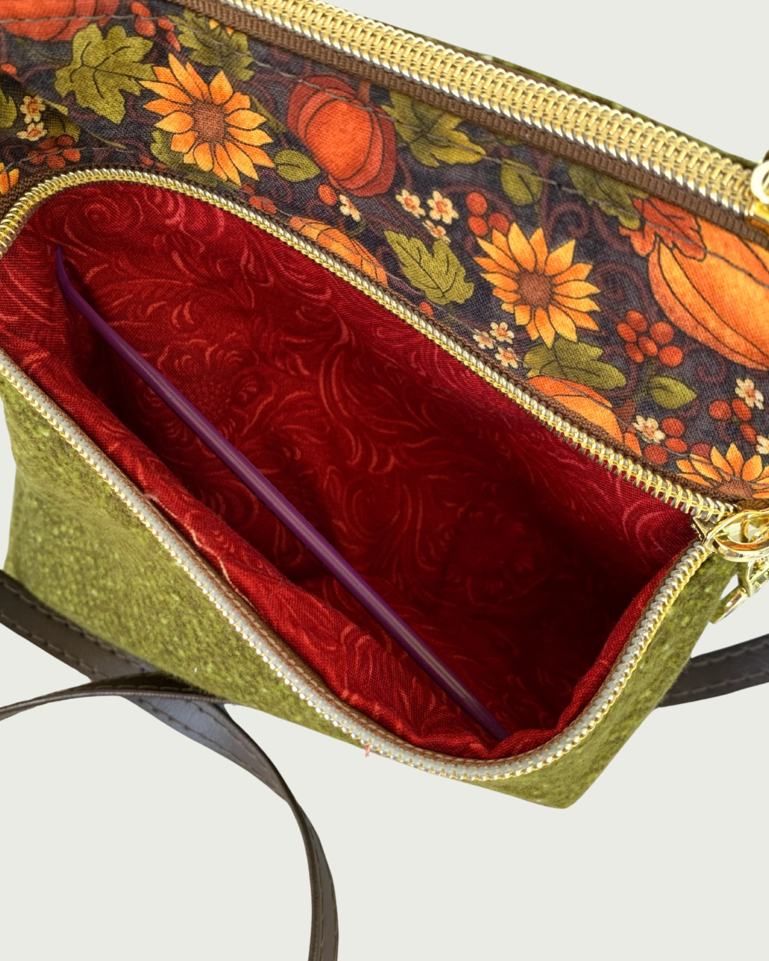 Handmade olive green crossbody bag closeup view of a textured fabric body, and a pumpkin and sunflower patterned accent. A gold-tone zippered pocket is open exposing a rich, rust colored cotton lining. A top zipper closure and a thin, coffee. adjustable and detachable shoulder strap can be partially seen.