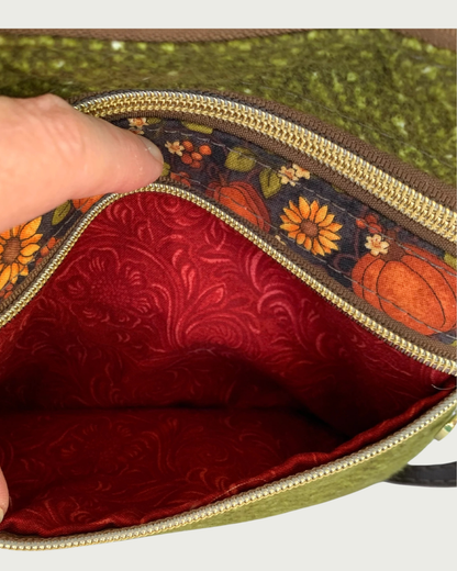 Handmade olive green crossbody bag closeup view of a textured fabric body, and a pumpkin and sunflower patterned accent. The bottom gold-tone zippered pocket is open exposing a rich, rust colored cotton lining. Another closed zippered pocket is shown above the open on.
