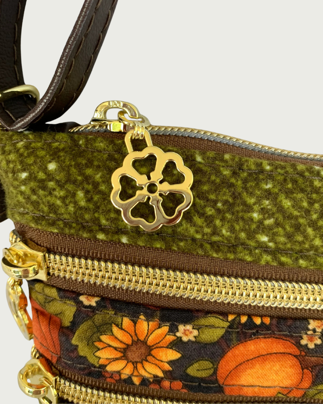 Handmade olive green crossbody bag closeup view of a textured fabric body, and a pumpkin and sunflower patterned accent. Two gold-tone zippered pockets and top zipper closure that shows a gold flower zipper pull. A thin, coffee adjustable and detachable shoulder strap can be partially seen.