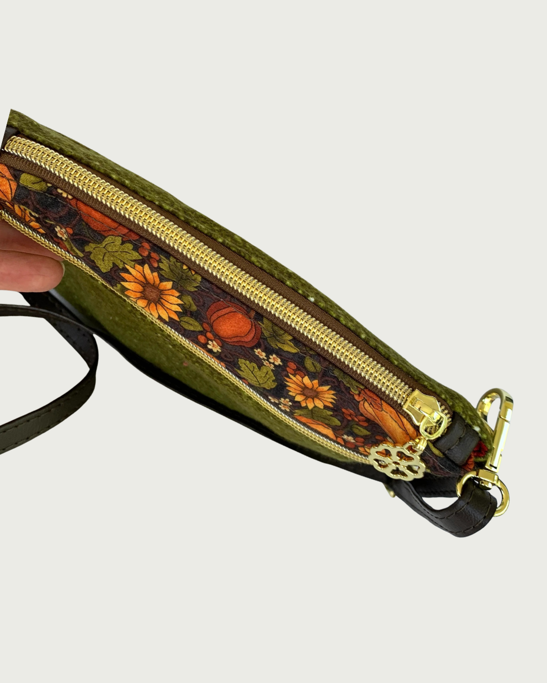 Handmade olive green crossbody bag being viewed looking down at the top. It has a textured fabric body, a zippered pocket is seen with a pumpkin and sunflower patterned band between it and the top zipper closure, gold-tone hardware including a flower zipper pull. A thin, coffee adjustable and detachable shoulder strap can be partially seen.