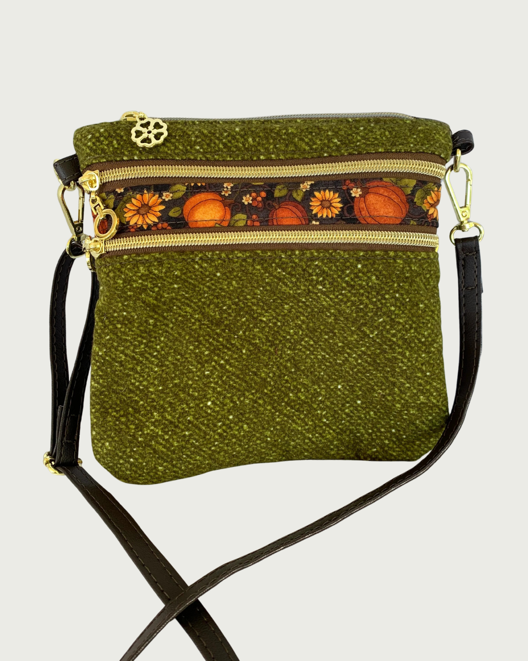 Handmade olive green crossbody bag with a textured fabric body, two zippered pockets featuring a pumpkin and sunflower patterned band between them, gold-tone hardware, a top zippered closure, and a thin, coffee adjustable and detachable shoulder strap.