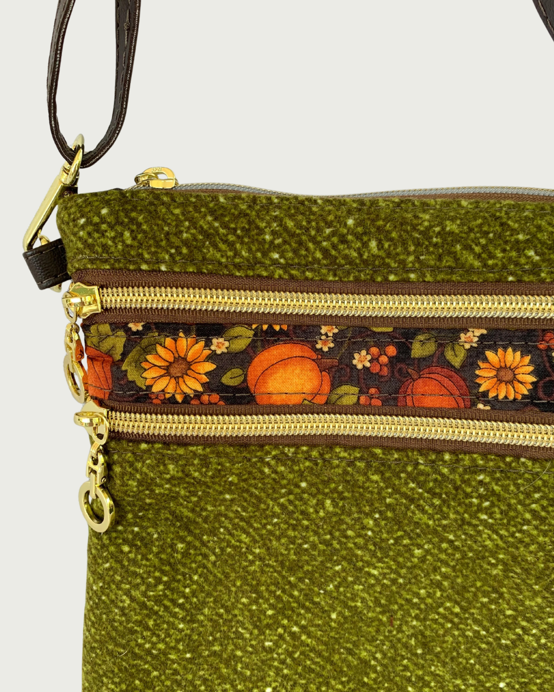 Handmade olive green crossbody bag closeup view with a textured fabric body, two zippered pockets featuring a pumpkin and sunflower patterned band between them, gold-tone hardware, a top zippered closure, and a thin, coffee adjustable and detachable shoulder strap.