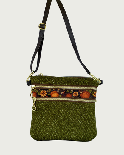 Handmade olive green crossbody bag with a textured fabric body, two zippered pockets featuring a pumpkin and sunflower patterned band between them, gold-tone hardware, a top zippered closure, hanging by a thin, coffee adjustable and detachable shoulder strap.