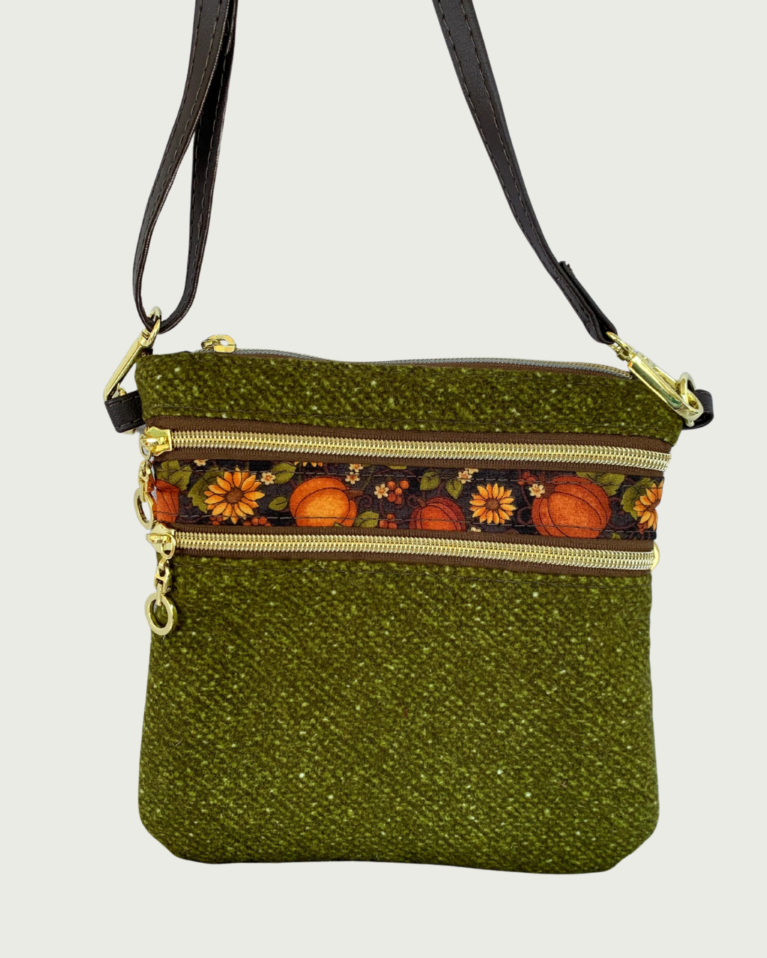 Handmade olive green crossbody bag with a textured fabric body, two zippered pockets featuring a pumpkin and sunflower patterned band between them, gold-tone hardware, a top zippered closure, hanging by a thin, coffee adjustable and detachable shoulder strap.