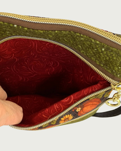 Handmade olive green crossbody bag closeup view of a textured fabric body, and a pumpkin and sunflower patterned accent. The top gold-tone zippered pocket is open exposing a rich, rust colored cotton lining. A top zipper closure, another zippered pocket, and a thin, coffee. adjustable and detachable shoulder strap can be partially seen.