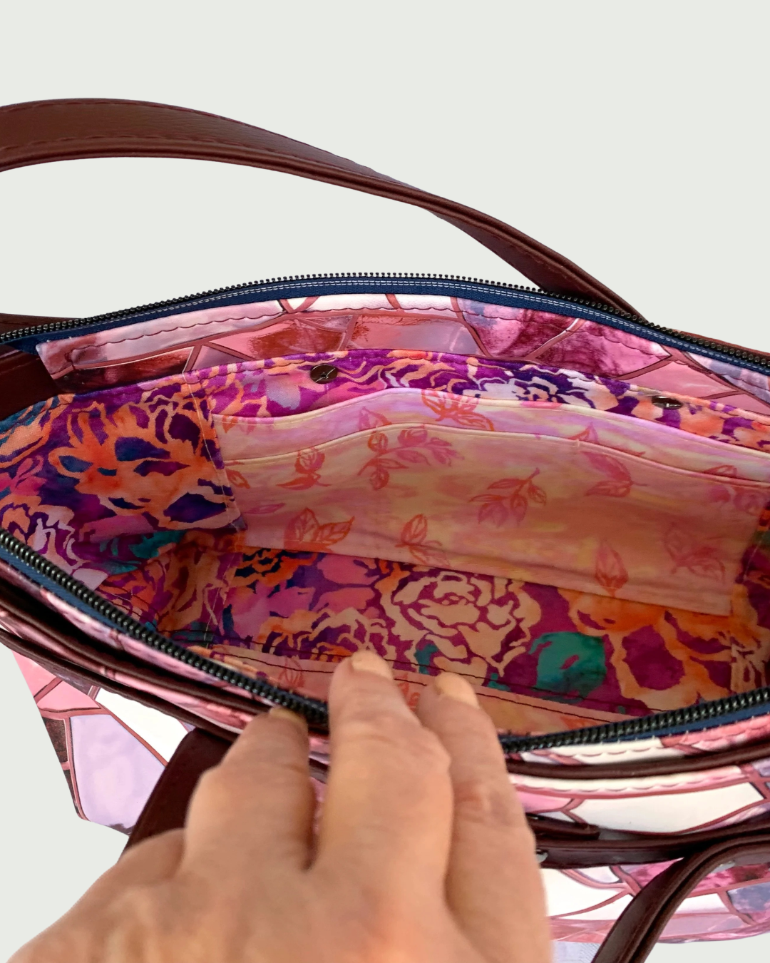 Handmade East-West style bag made from vinyl in a print of fracture glass in colors of pink, rose, maroon, and blue. Two handles made from maroon marine vinyl are attached, front and back, to a wrap-around strap detail. The view is of the open interior of the bag which is made of cotton woven in two floral prints of pink, purple and green. A combination of a slip pocket and a three-cardslot panel is sewn into the side. The top zipper is navy tape with gunmetal teeth. 