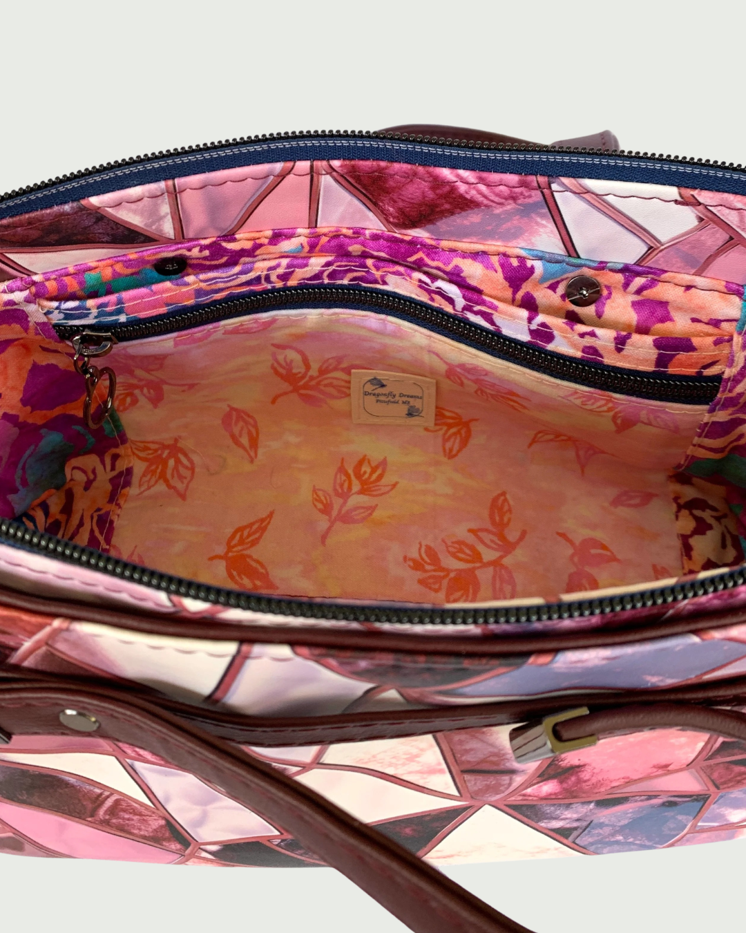 Handmade East-West style bag made from vinyl in a print of fracture glass in colors of pink, rose, maroon, and blue. Two handles made from maroon marine vinyl are attached, front and back, to a wrap-around strap detail. The view is of the open interior of the bag which is made of cotton woven in two floral prints of pink, purple and green. A combination of a slip pocket and a zipper pocket panel is on the side with a Dragonfly Dreams label sewn into the pocket front. The top zipper is navy tape with gunmeta
