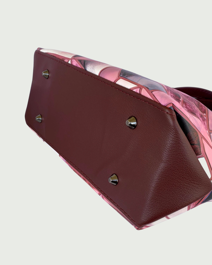 Handmade East-West style satchel in a close up view of the bottom. The bag is made from vinyl in a print of fracture glass in colors of pink, rose, maroon, and blue. The bottom is maroon faux leather maroon and a surplus piece on the side is shaped into a triangle that is attached to the side of the bag. There are four bucket-shaped gunmetal bag feet.