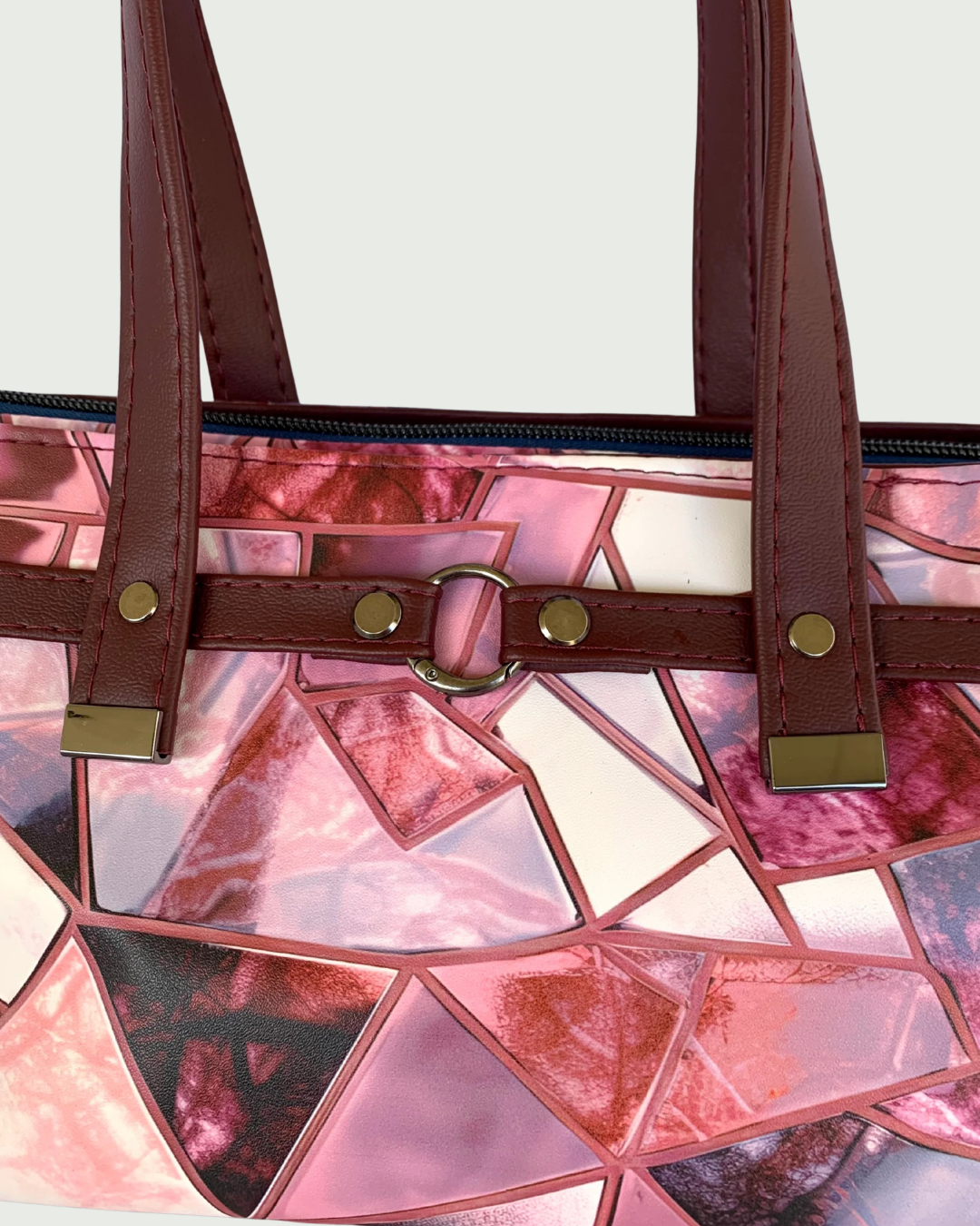 Handmade East-West style satchel in a close up view of the front. The bag is made from vinyl in a print of fracture glass in colors of pink, rose, maroon, and blue. Two handles made from maroon marine vinyl are attached to a wrap-around strap detail that is held together with a caribiner ring in the front. All hardware is a shiny gunmetal. 