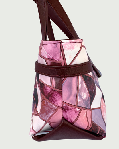 Handmade East-West style satchel viewed from the side. The bag is made from vinyl in a print of fracture glass in colors of pink, rose, maroon, and blue with maroon faux leather details including a wrap-around strap detail and a triangular bottom extension.  