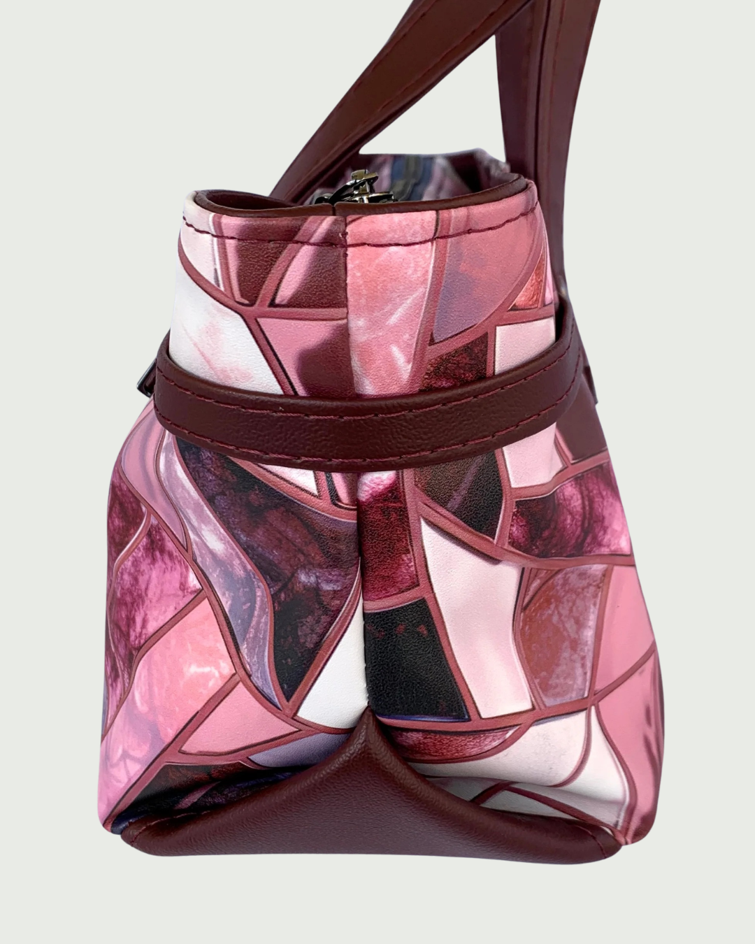 Handmade East-West style satchel viewed from the side. The bag is made from vinyl in a print of fracture glass in colors of pink, rose, maroon, and blue with maroon faux leather details including a wrap-around strap detail and a triangular bottom extension.  