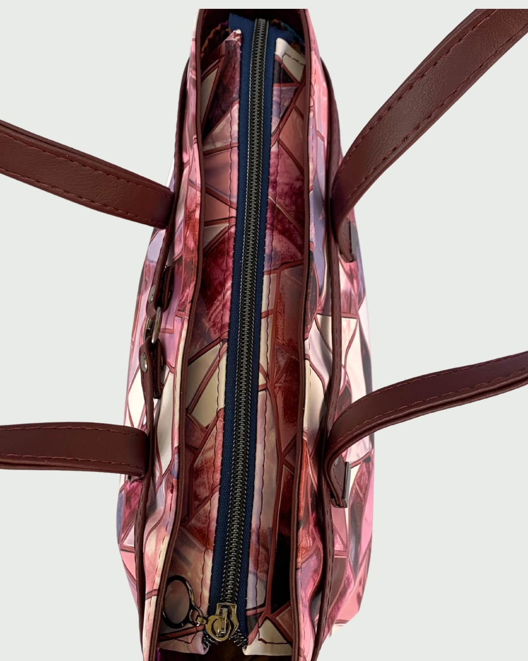 Handmade East-West style satchel in a close up view from above. The bag is made from vinyl in a print of fracture glass in colors of pink, rose, maroon, and blue. Two handles made from maroon marine vinyl are attached to a wrap-around strap detail that is held together with a caribiner ring in the front. The handles are separated to view the recessed zipper of navy zipper tape and an open circle gunmetal zipper pull.