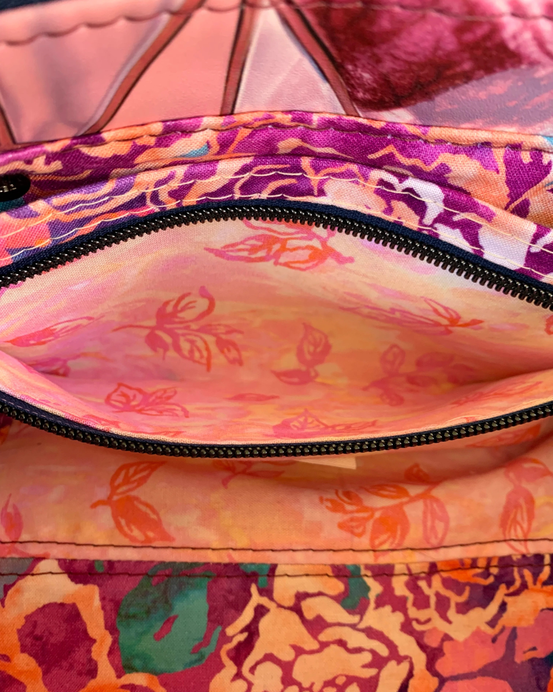 Handmade East-West style bag interior which is made of cotton woven in two floral prints of pink, purple and green. A combination of a slip pocket and a zipper pocket panel is sewn into the side. The zipper is open displaying the pocket interior. The zipper is navy tape with gunmetal teeth.