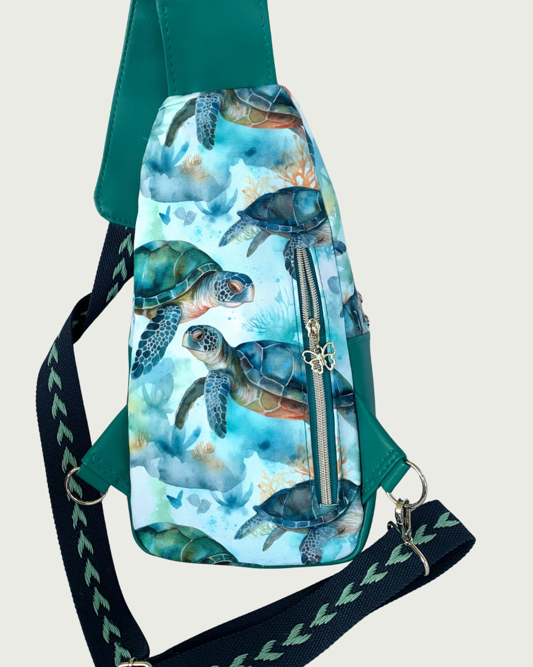 Back view of handcrafted sling bag with a print of sweet sea turtles in teal, aqua, green, brown, and white. Water-resistant canvas bag with an adjustable navy and aqua wide cotton webbing strap also repositional for left and right handed wear. All hardware is nickel including the zipper teeth on teal zipper tape that is positioned vertically for a concealed pocket and a sweet butterfly pull.