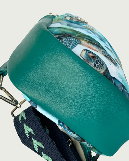 The bottom view of a handcrafted sling bag of water-resistant canvas in teal, aqua, and brown. The bottom is also part of the gusset and it is made from teal faux leather. An adjustable navy and aqua wide cotton webbing strap is shown. All hardware is nickel.