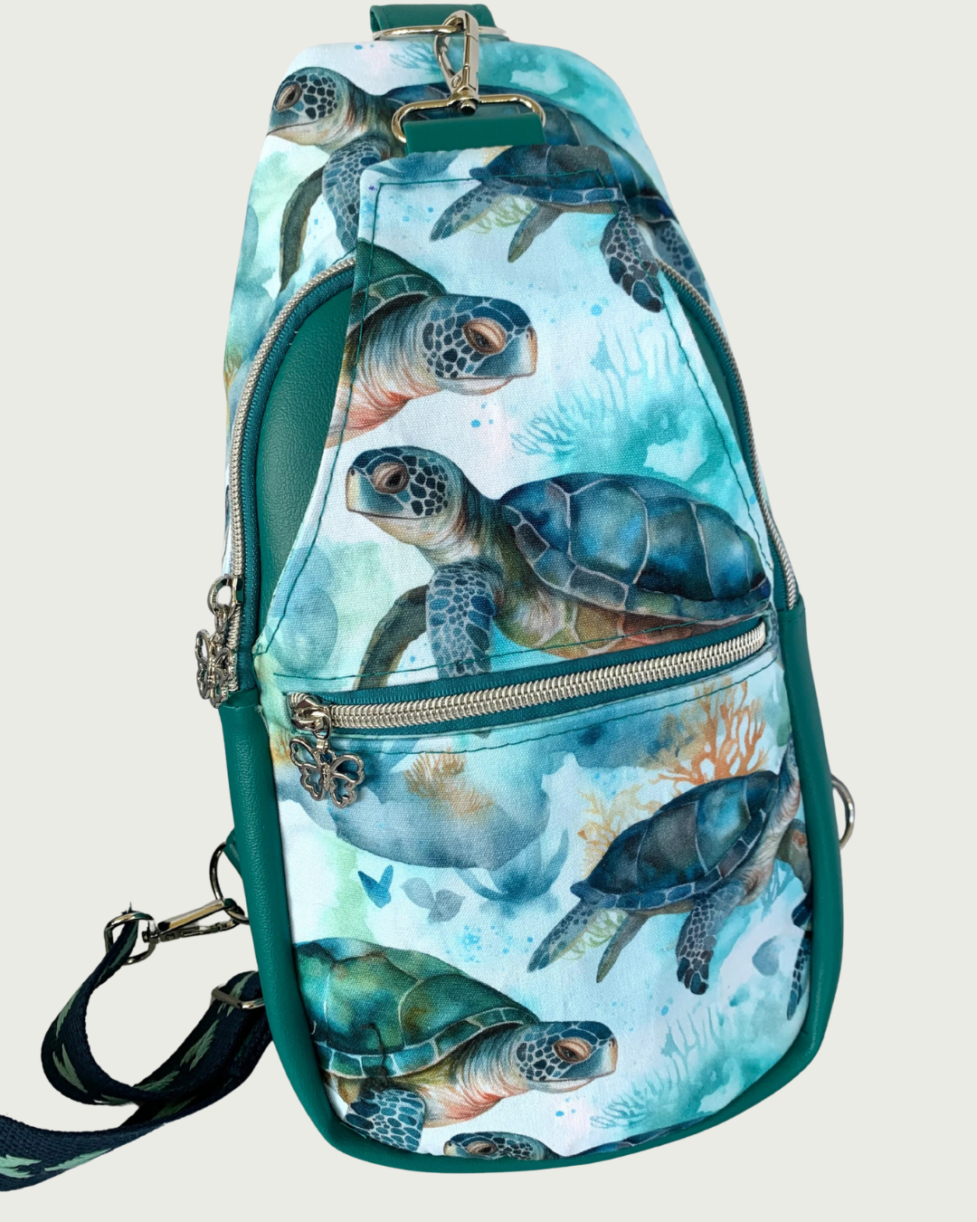 Handcrafted sling bag with a print of sweet sea turtles in teal, aqua, green, brown, and white. Water-resistant canvas bag with teal faux leather accents with an adjustable navy and aqua wide cotton webbing strap also repositional for left and right handed wear. All hardware is nickel including the zipper teeth on teal zipper tape and the butterfly zipper pulls. A front zipper pocket is shown as well as a front panel that unhooks with a swivel held onto a d-ring. Behind that is a curved zippered main compar