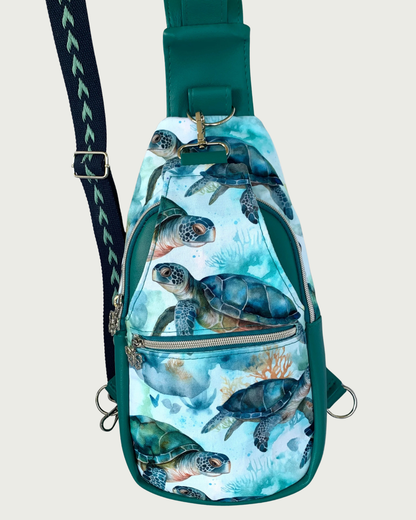 Handcrafted sling bag with a print of sweet sea turtles in teal, aqua, green, brown, and white. Water-resistant canvas bag with teal faux leather accents with an adjustable navy and aqua wide cotton webbing strap also repositional for left and right handed wear. All hardware is nickel including the zipper teeth on black zipper tape. A front zipper pocket is shown as well as a front panel that unhooks with a swivel held onto a d-ring. Behind that is a curved zippered main compartment of teal vinyl.