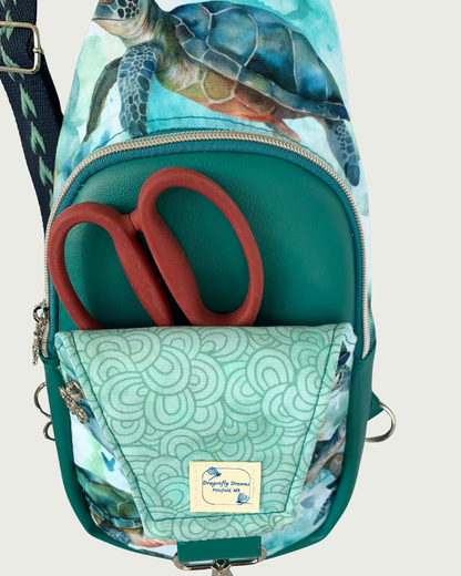 Handcrafted sling bag with a print of sweet sea turtles in teal, aqua, green, brown, and white. Water-resistant canvas bag with teal faux leather accents and a wide, teal and aqual adjustable and repositional cotton webbing strap. A pair of red-handled scissors is in the pocket space between an open panel of aqua lining and the main compartment of teal faux leather vinyl. The compartment is zipped closed with teal zipper tape and nickel teeth. A Dragonfly Dreams label is sewn into the lining.