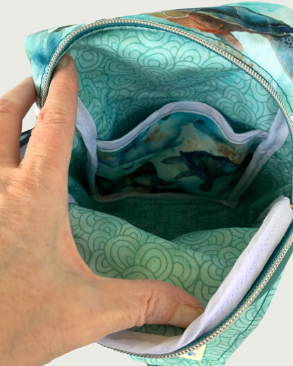 Handcrafted sling bag made of water-resistant canvas. A hand is holding open the unzipped main compartment revealing an aqua lining, a slip pocket with it’s face made of a teal sea turtle print, and white binding on the seams.