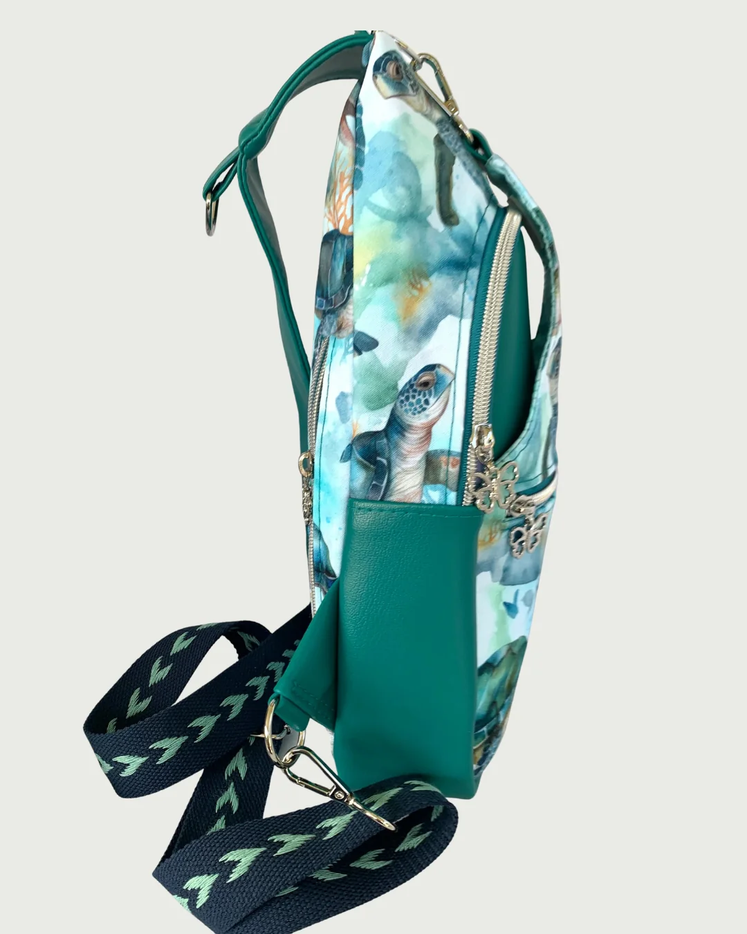 Handcrafted sling bag standing sideways of water-resistant canvas with a print of sweet sea turtles in teal, aqua, green, brown, and white. Teal faux leather accents with an adjustable navy and aqua wide cotton webbing strap. All hardware is nickel including the zipper teeth on teal zipper tape on the front pocket as well as the main compartment. A front panel that has the pocket unhooks with a swivel held onto a d-ring. Behind that is a curved zippered main compartment made of the teal vinyl. The zipper pu